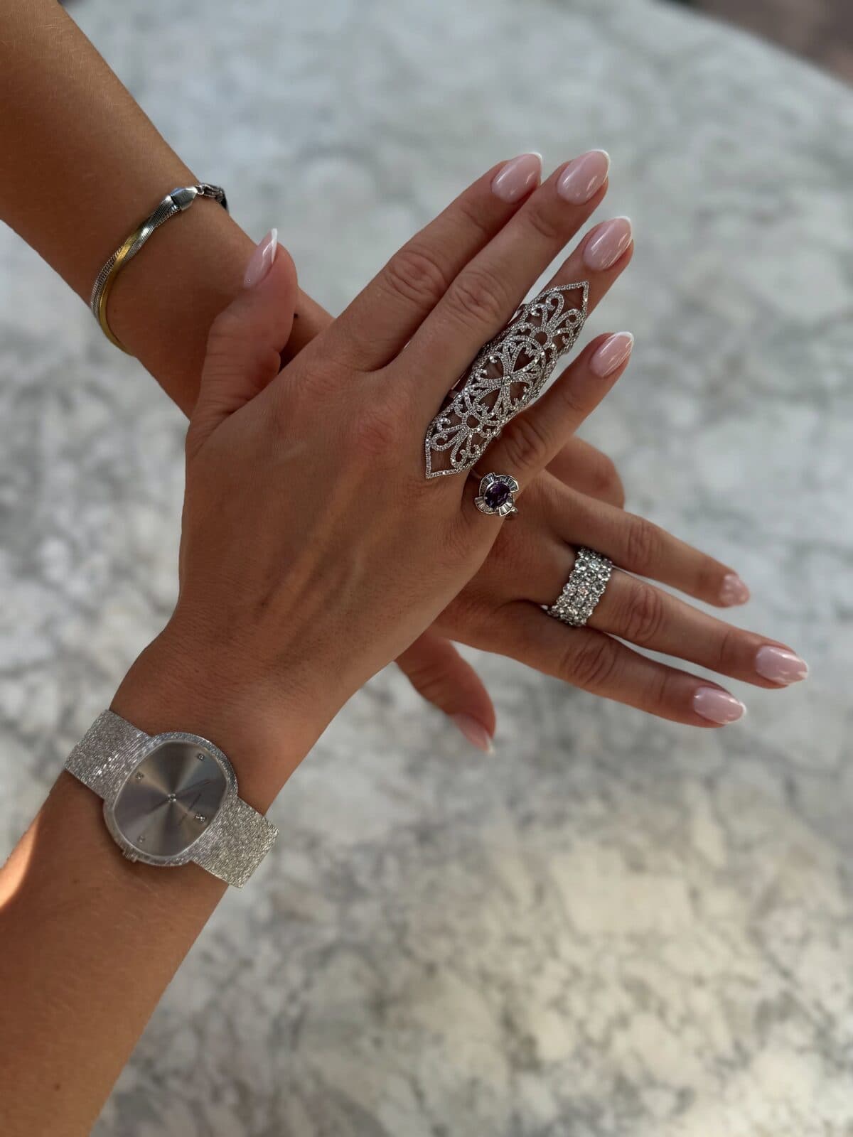 Manos cruzadas con anillos, una pulsera y un reloj sobre mármol claro.